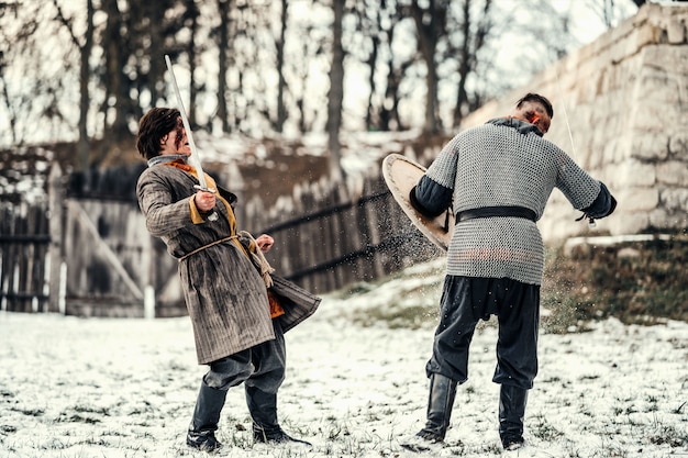 Dos guerreros antiguos en armadura con armas luchando con espadas en la nieve.