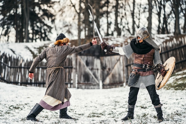 Dos guerreros antiguos en armadura con armas luchando con espadas en la nieve.