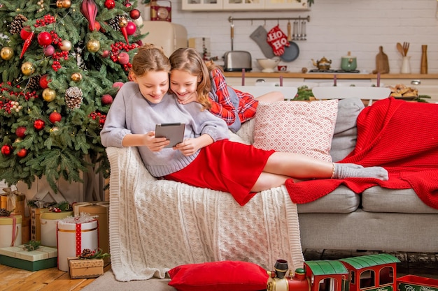 Dos hermanas alegres están sentadas en casa cerca del árbol de Navidad y mirando el monitor de la tableta