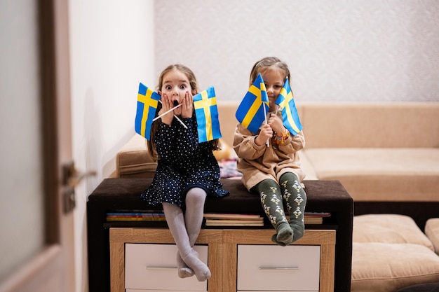 Dos hermanas están sentadas en un sofá en casa con banderas suecas en las manos Suecia niños niñas con bandera