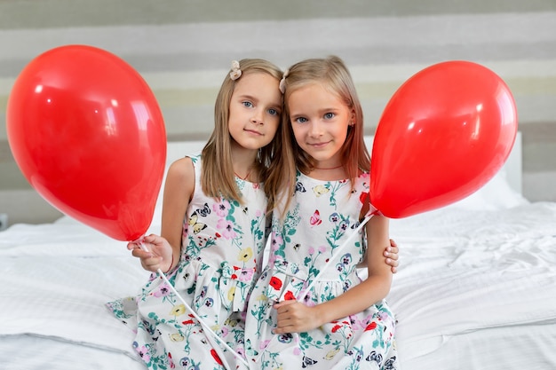 Dos hermanas gemelas con hermosos vestidos están sentadas en una cama en un hotel con globos en sus manos.