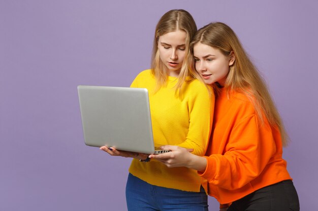 Dos hermanas gemelas rubias bastante jóvenes niñas en ropa de colores vivos sosteniendo, usando una computadora portátil aislada en la pared azul violeta pastel. Concepto de estilo de vida familiar de personas.