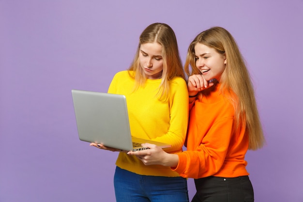 Dos hermanas gemelas rubias bastante jóvenes niñas en ropa de colores vivos sosteniendo, usando una computadora portátil aislada en la pared azul violeta pastel. Concepto de estilo de vida familiar de personas.