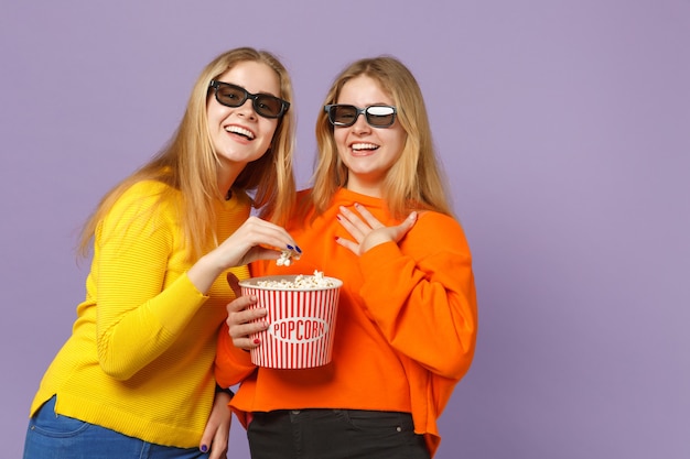 Dos hermanas gemelas rubias jóvenes riendo a niñas en gafas imax 3d viendo una película, sostienen palomitas de maíz aisladas en la pared azul violeta pastel. Concepto de estilo de vida familiar de personas.