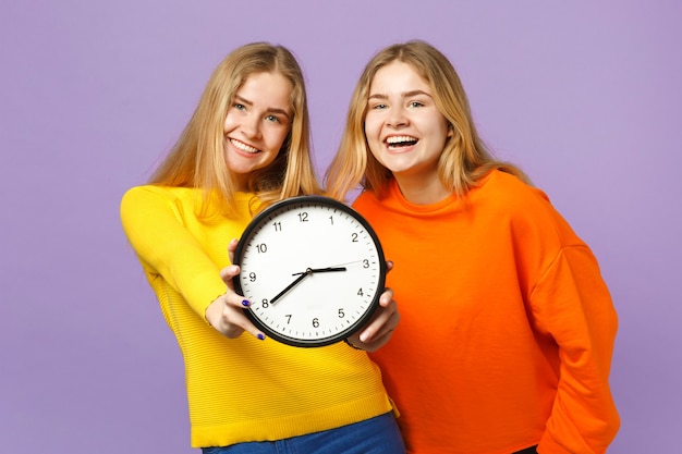 Dos hermanas gemelas rubias jóvenes riendo niñas en ropa de colores vivos sostienen el reloj redondo aislado en la pared azul violeta pastel. Concepto de estilo de vida familiar de personas.