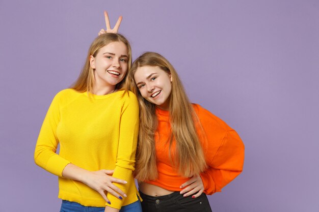 Dos hermanas gemelas rubias jóvenes sonrientes a niñas en ropa de colores vivos divirtiéndose, aisladas en la pared azul violeta pastel. Concepto de estilo de vida familiar de personas.