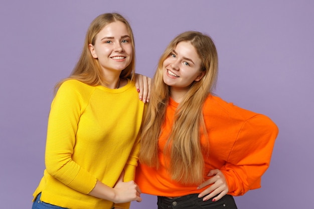 Dos hermanas gemelas rubias jóvenes sonrientes a niñas en ropa de colores vivos de pie, aisladas en la pared azul violeta pastel. Concepto de estilo de vida familiar de personas.