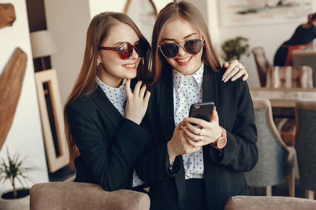 dos hermanas jóvenes y con estilo de pie en una cafetería y usando el teléfono