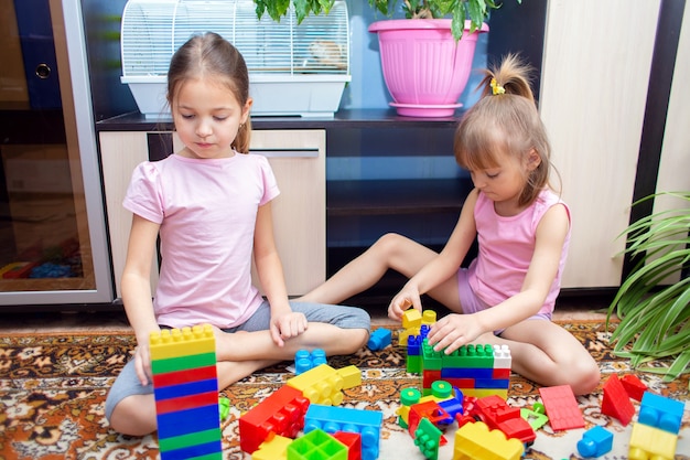 Dos hermanas niñas están sentadas en casa y juegan un juego de construcción de plástico. Los niños construyen juguetes a partir de bloques de colores.