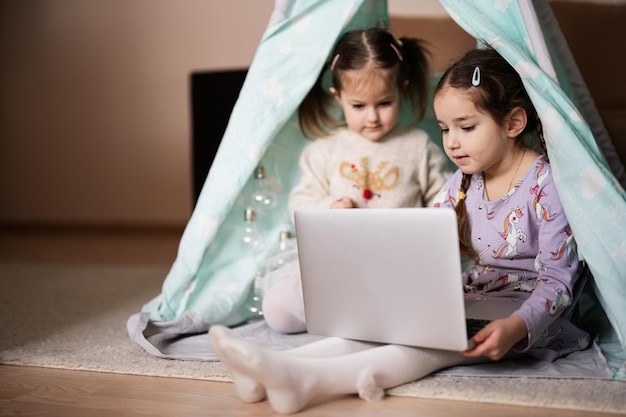 Dos hermanas niñas mirando en la computadora portátil en la tienda wigwam Tecnología y concepto de hogar