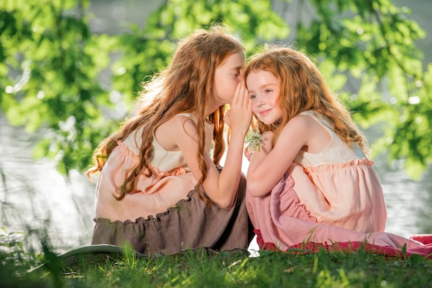 Dos hermanas pelirrojas con vestidos largos de lino descansan en el lago en el parque en un día soleado de verano.