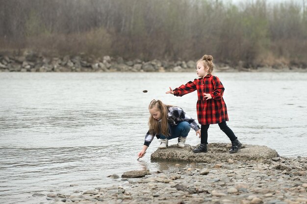 dos hermanas vestidas con ropa a cuadros juegan alegremente junto al río y arrojan piedras al agua