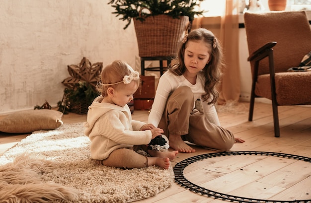 Dos hermanitas están sentadas en la alfombra y jugando con un tren de juguete cerca del árbol de Navidad en la habitación.