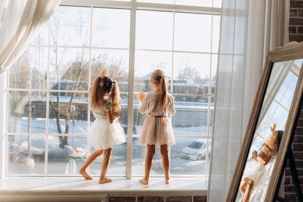 Dos hermanitas vestidas con hermosos vestidos se paran en el alféizar de la ventana junto al espejo.