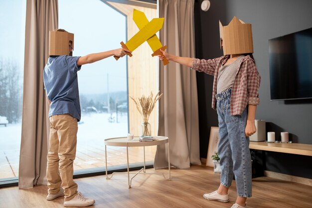 Dos hermanitos en ropa casual y cascos de cartón jugando con espadas de juguete contra una ventana grande con cortinas beige en la sala de estar