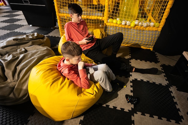 Dos hermanos jugando a la consola de videojuegos sentados en un puf amarillo en el centro de juegos para niños