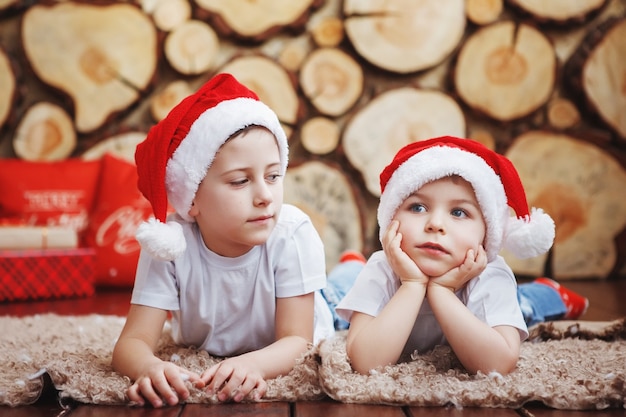 Dos hermanos varones con sombreros de Navidad están mintiendo y riendo en la alfombra cerca del árbol de Año Nuevo en el fondo de los cortes de árboles.