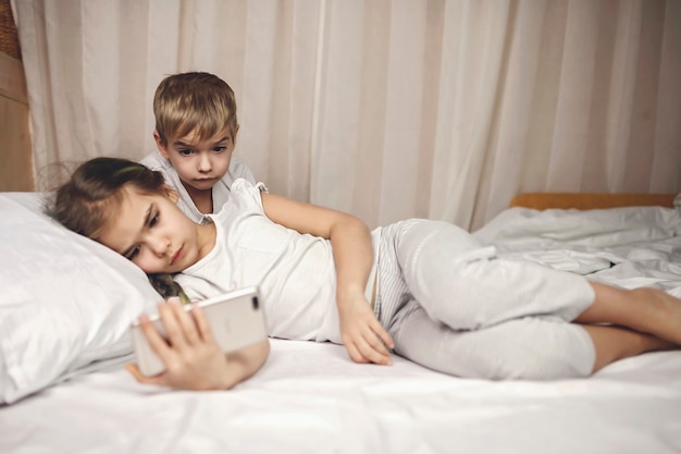 Dos hermanos viendo videos en dispositivos electrónicos móviles en la cama, adicción a los aparatos