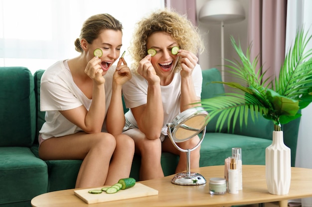 Dos hermosas y alegres chicas aplicando rodajas de pepino para la piel debajo de los ojos en casa