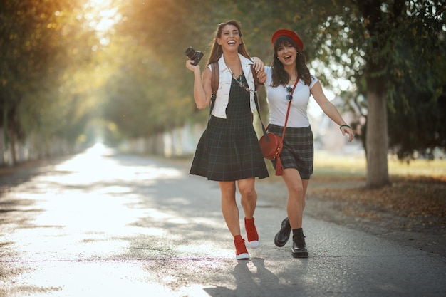 Foto dos hermosas y alegres mujeres jóvenes con mochilas en la espalda y cámara fotográfica caminan por la soleada avenida otoñal.