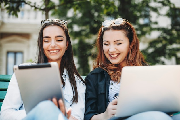 Dos hermosas amigas caucásicas jóvenes sentados en un banco con una computadora portátil y una tableta mirando en la tableta riendo.