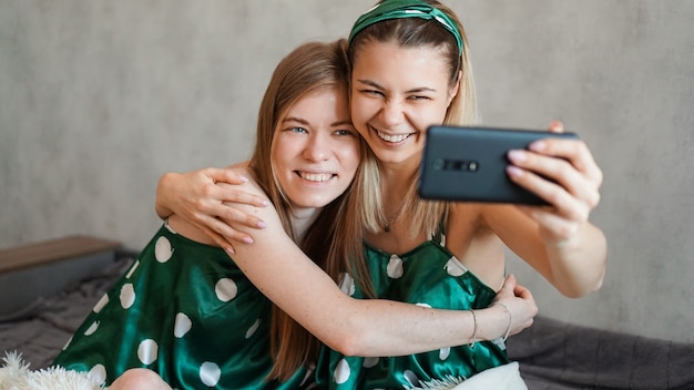 Dos hermosas amigas felices abrazándose y tomando selfies con el teléfono inteligente en la fiesta de pijamas