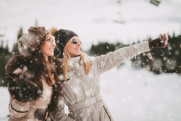 Dos hermosas amigas felices disfrutando de las vacaciones de invierno. Están dando un paseo mientras está nevando y mirando hacia otro lado.