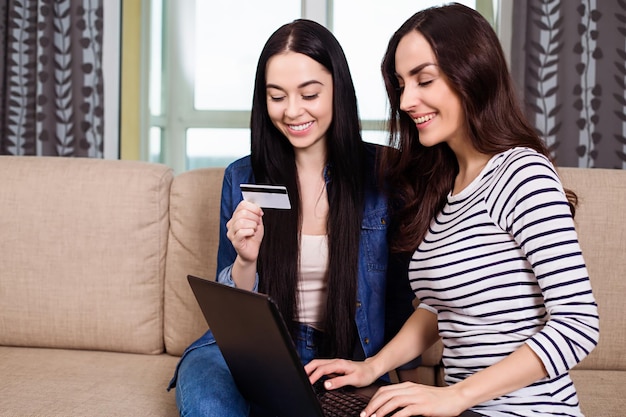 Dos hermosas amigas o hermanas sonrientes hacen un pedido en línea o compran en Internet con la ayuda de una tarjeta bancaria