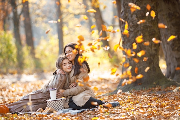 Dos hermosas amigas pasando tiempo en una manta de picnic en el césped. Dos jóvenes hermanas sonrientes haciendo picnic comiendo croissant en el parque de otoño. Chicas morenas y rubias con abrigos.