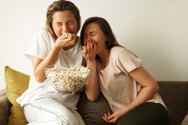 Dos hermosas amigas viendo programas de televisión y comiendo palomitas de maíz en casa