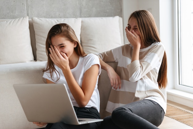 Dos hermosas chicas adolescentes alegres que usan la computadora portátil mientras están sentados en casa