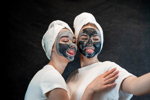 Dos hermosas chicas aplicando máscara negra de arcilla aisladas en negro Hogar cuidar de la piel de la cara Estilo de vida