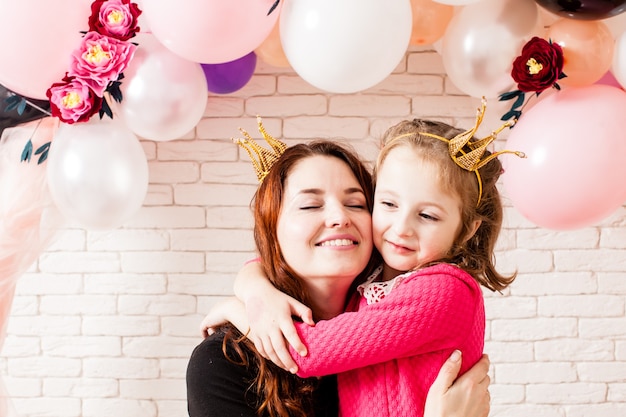 Dos hermosas chicas con coronas debajo de globos de cumpleaños y decoraciones de arco de flores de papel. Photozone infantil para celebración. Mamá con hija