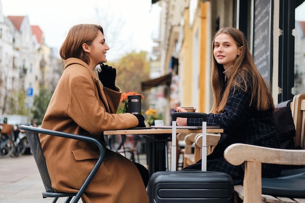 Dos hermosas chicas con estilo felizmente bebiendo café juntas mientras están sentadas en un café de la calle con una maleta