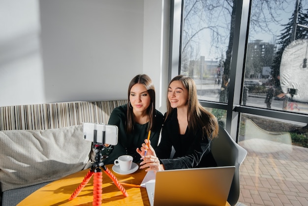 Dos hermosas chicas jóvenes están sentadas en un café, grabando blogs de video y comunicándose en las redes sociales.