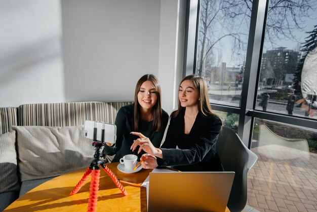 Dos hermosas chicas jóvenes están sentadas en un café, grabando blogs de video y comunicándose en las redes sociales.