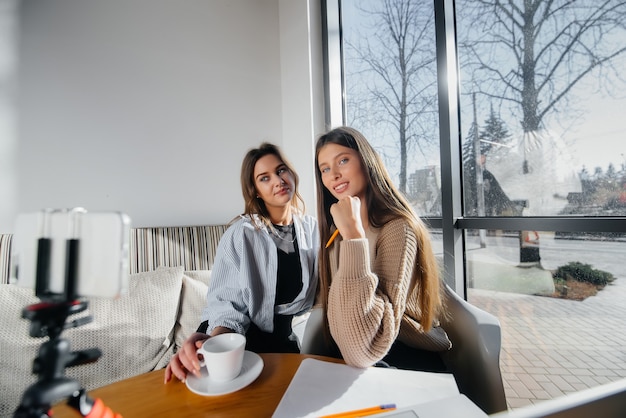 Dos hermosas chicas jóvenes están sentadas en un café, grabando blogs de video y comunicándose en las redes sociales.