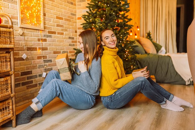 Dos hermosas chicas jóvenes felices alegres novia dan regalos de Navidad en el fondo de un árbol de año nuevo en casa