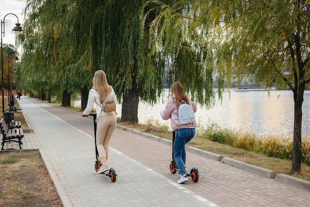 Dos hermosas chicas jóvenes en máscaras andan en patinetes eléctricos en el parque en un cálido día de otoño