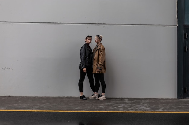 Dos hermosas chicas jóvenes con un traje de otoño de moda con una chaqueta de cuero vintage y jeans negros están de pie cerca de una pared gris minimalista en la calle