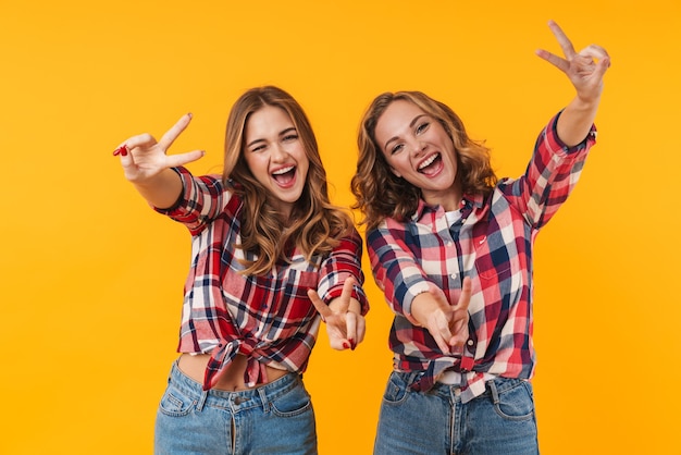 Foto dos hermosas chicas jóvenes vistiendo camisa a cuadros sonriendo y gesticulando el signo de la paz aislado
