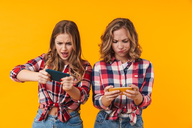 Dos hermosas chicas jóvenes vistiendo camisetas a cuadros divirtiéndose y jugando videojuegos en teléfonos móviles aislados