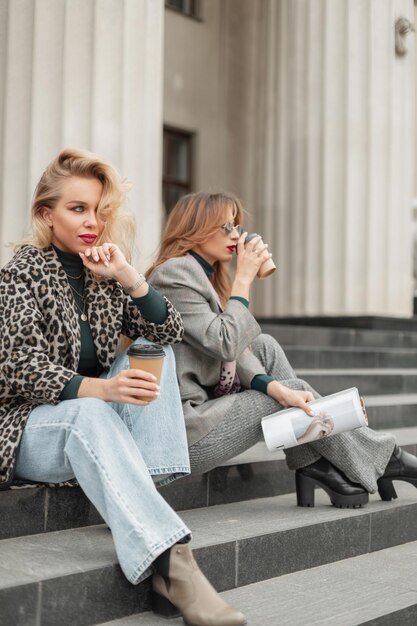 Dos hermosas chicas modelo de moda con ropa elegante se sientan en los escalones de la ciudad y beben café