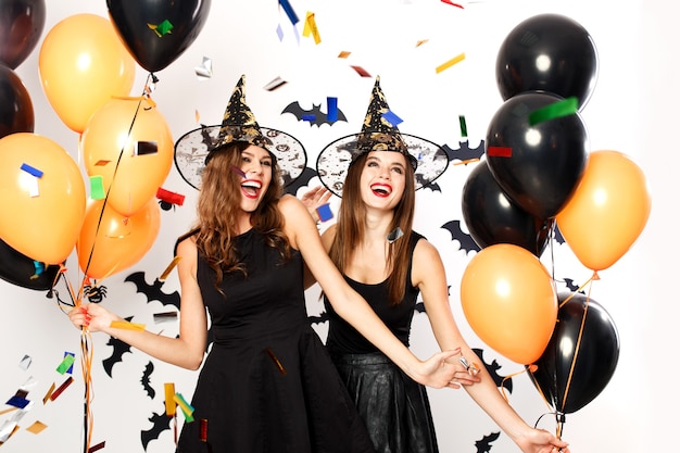 Dos hermosas chicas con vestidos negros y sombreros de bruja se divierten con globos negros y naranjas y confeti.Fiesta de Halloween.