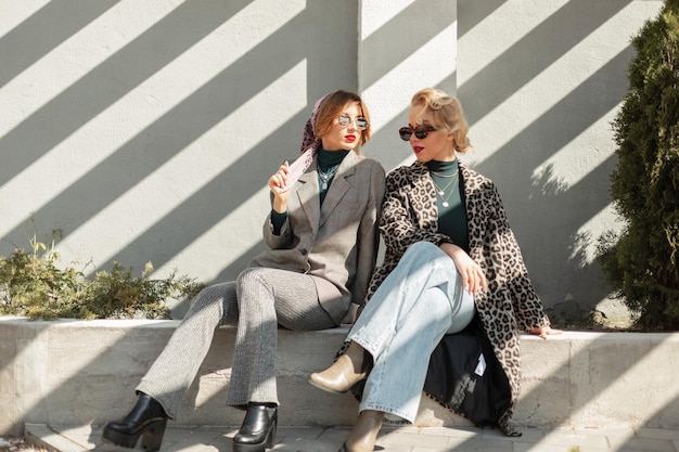 Dos hermosas y elegantes novias de la modelo vestidas a la moda con abrigos de leopardo, pantalones vaqueros, un traje y zapatos, se sientan en la ciudad con la luz del sol. Estilo vintage urbano y ropa de moda.