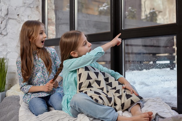 Dos hermosas hermanas caucásicas sentadas en el alféizar de la ventana, vieron algo interesante en la ventana e intentaron verlo.