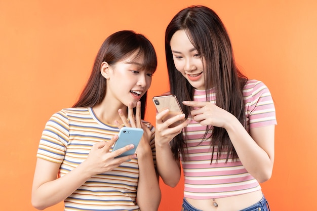 Dos hermosas jóvenes asiáticas están usando teléfonos móviles en la pared naranja