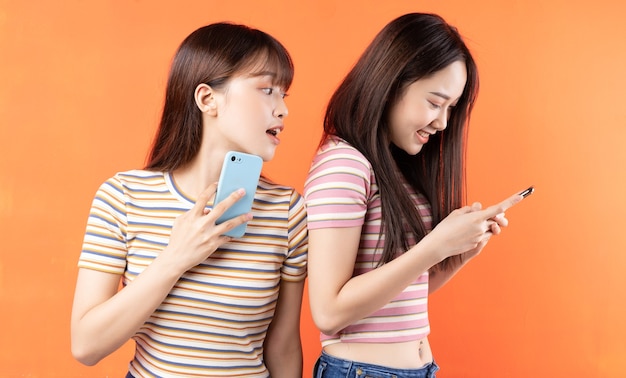 Dos hermosas jóvenes asiáticas están usando teléfonos móviles en la pared naranja