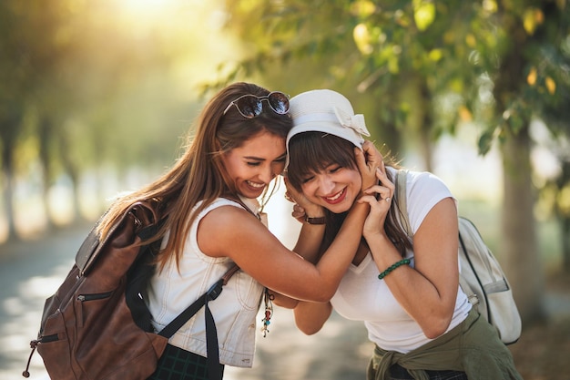 Dos hermosas jóvenes sonrientes con mochilas en la espalda caminan por la avenida soleada de otoño, abrazándose y hablando entre ellas.