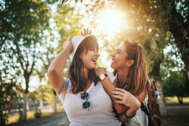 Dos hermosas jóvenes sonrientes con mochilas en la espalda caminan por la avenida soleada de otoño, abrazándose y hablando entre ellas.